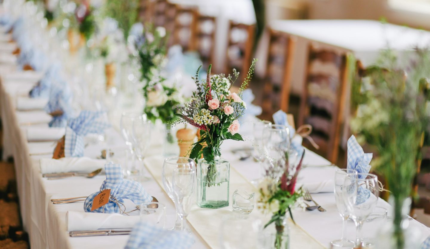 Mesa de festa clara, com guardanapos azuis e pequenos arranjos de flores